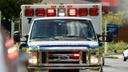 An ambulance seen head on with its lights on in the daylight.