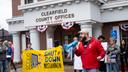 A man in a red jacket speaks into a megaphone in front of the Clearfield County Offices building. Next to him are protesters holding a yellow sign that says “Shut Down Moshannon.”