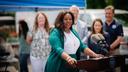 Sally Zubairu-Cofield speaks at an outdoor news conference from behind a podium with two microphones, with several people and a blue canopy behind her.