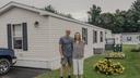 A man and woman stand in front of a mobile home with yellow flowers next to them.