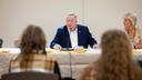 Aging Secretary Jason Kavulich speaks into a microphone during a meeting, with attendees in the foreground and a woman seated beside him.