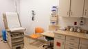 A clinical examination room with a brown examination table, orange chair, medical equipment on the wall, and a cabinet holding medical supplies.