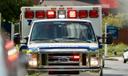 An ambulance seen head on with its lights on in the daylight.