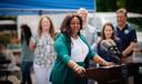 Sally Zubairu-Cofield speaks at an outdoor news conference from behind a podium with two microphones, with several people and a blue canopy behind her.