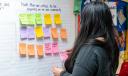 Listening session participant looking at a wall with sticky notes holding a pink sticky note with the words road, project, updates written on it.