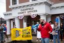 A man in a red jacket speaks into a megaphone in front of the Clearfield County Offices building. Next to him are protesters holding a yellow sign that says “Shut Down Moshannon.”