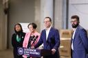 News conference featuring Governor Josh Shapiro at a podium, flanked by three individuals in a spacious industrial setting with cardboard boxes in the background.