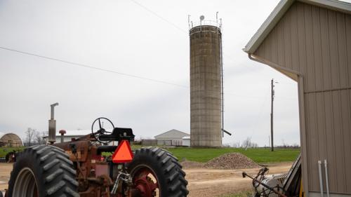 A tractor in the foreground and a radios and a microwave dish providing internet service installed atop a farm silo in the background.