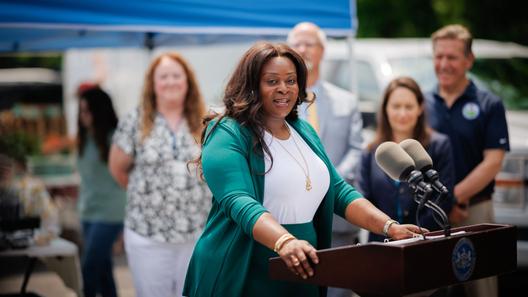 Sally Zubairu-Cofield speaks at an outdoor news conference from behind a podium with two microphones, with several people and a blue canopy behind her.