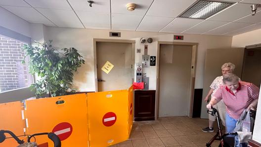 Two women, one leaning on a walker, stand in a hall near two elevators. One elevator with an out of order sign on the door has four barriers in front of it.