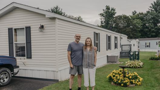 A man and woman stand in front of a mobile home with yellow flowers next to them.