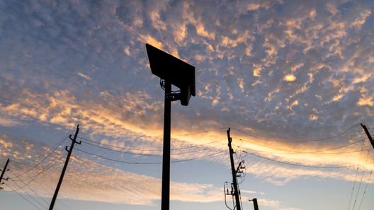 Silhouetted Flock Safety license plate reader against an orange-and-blue sunset sky, with utility poles and power lines below.