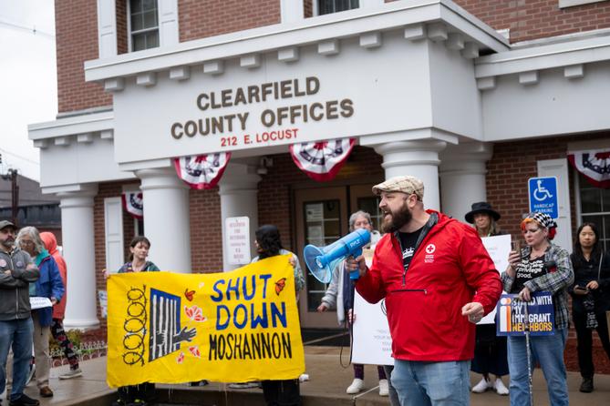 A man in a red jacket speaks into a megaphone in front of the Clearfield County Offices building. Next to him are protesters holding a yellow sign that says “Shut Down Moshannon.” A man in a red jacket speaks into a megaphone in front of the Clearfield County Offices building. Next to him are protesters holding a yellow sign that says “Shut Down Moshannon.”