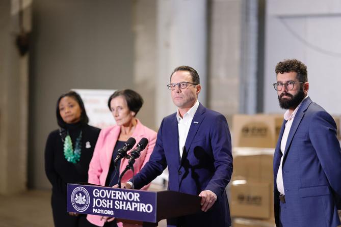 News conference featuring Governor Josh Shapiro at a podium, flanked by three individuals in a spacious industrial setting with cardboard boxes in the background. News conference featuring Governor Josh Shapiro at a podium, flanked by three individuals in a spacious industrial setting with cardboard boxes in the background.