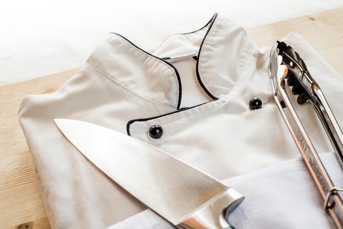 A stock image of a chef's coat and tongs on a wooden board. A stock image of a chef's coat and tongs on a wooden board.