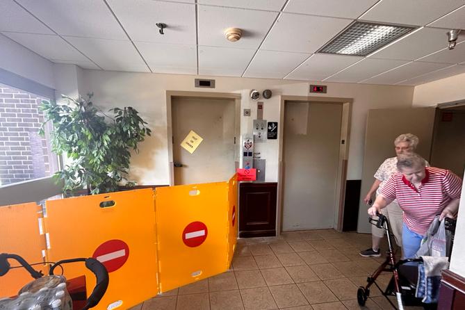 Two women, one leaning on a walker, stand in a hall near two elevators. One elevator with an out of order sign on the door has four barriers in front of it. Two women, one leaning on a walker, stand in a hall near two elevators. One elevator with an out of order sign on the door has four barriers in front of it.