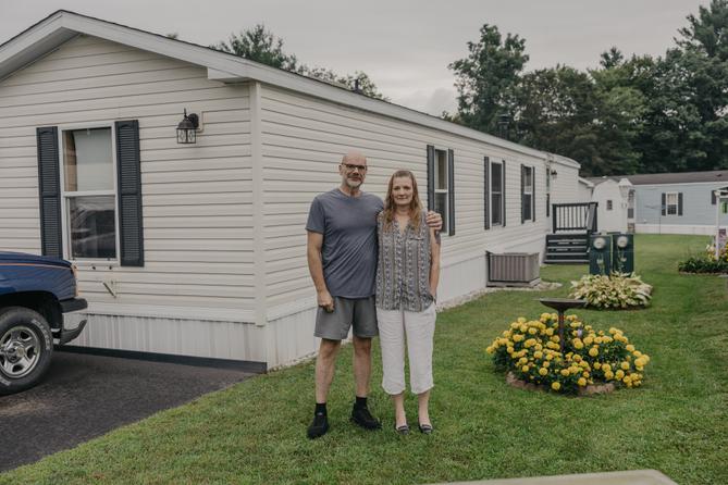 A man and woman stand in front of a mobile home with yellow flowers next to them. A man and woman stand in front of a mobile home with yellow flowers next to them.