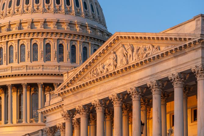 Close-up of the U.S. Capitol dome and an ornate portico illuminated by warm sunlight against a blue sky. Close-up of the U.S. Capitol dome and an ornate portico illuminated by warm sunlight against a blue sky.