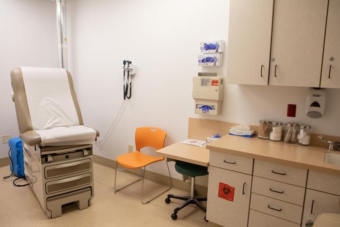 A clinical examination room with a brown examination table, orange chair, medical equipment on the wall, and a cabinet holding medical supplies. A clinical examination room with a brown examination table, orange chair, medical equipment on the wall, and a cabinet holding medical supplies.
