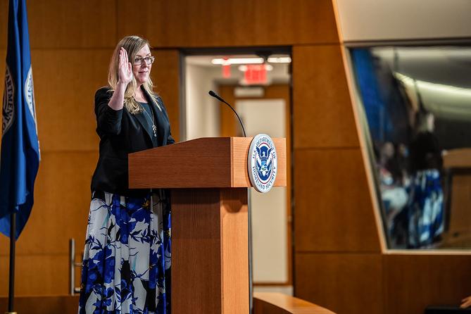 Jennifer Higgins, then-USCIS deputy director, swears in new citizens while raising her hand at a podium with the USCIS seal. Jennifer Higgins, then-USCIS deputy director, swears in new citizens while raising her hand at a podium with the USCIS seal.